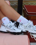 White sneakers with black soles and pink brand logo socks worn by a person sitting on a chair.